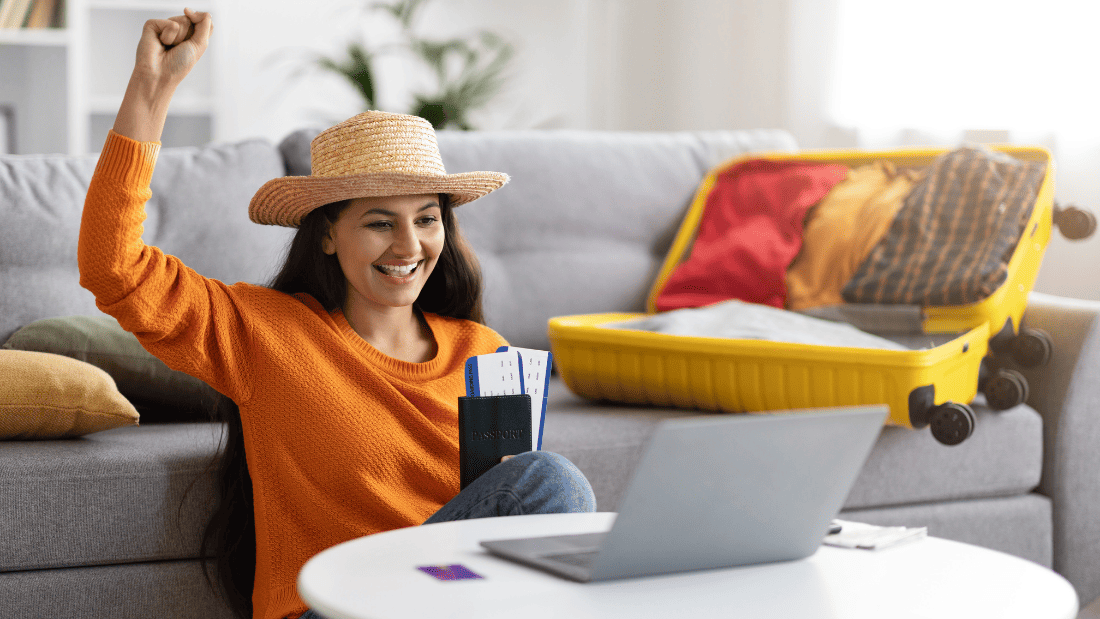 Jovem animada, olhando o notebook, com passagens aéreas nas mãos e uma mala de viagem ao fundo